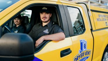 Two men in black uniforms with Dryer Vent Wizard logos sit in a bright yellow service truck.