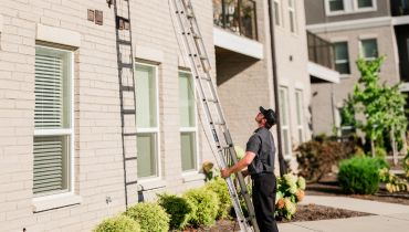 A man in a dark uniform and cap stands outside a light-colored brick building, looking up while holding a tall metal extension ladder against a wall.