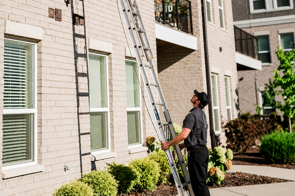A man in a dark uniform and cap stands outside a light-colored brick building, looking up while holding a tall metal extension ladder against a wall.