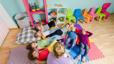 A teacher sits on a colorful foam mat reading to a group of seven diverse children lying on the floor in a bright classroom.