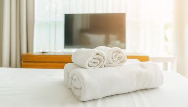 Two rolled white hand towels rest on a folded white bath towel on a bed, with a TV and sheer curtains in the background.