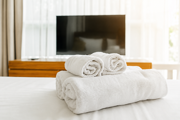 Two rolled white hand towels rest on a folded white bath towel on a bed, with a TV and sheer curtains in the background.