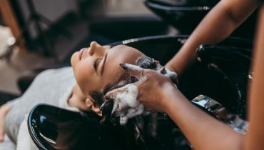 A person reclines at a salon sink while another person washes their hair, creating lather.